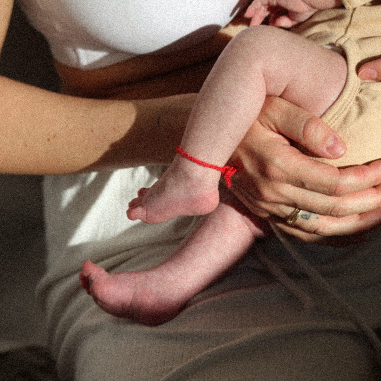 Eine Mutter hält ihr Neugeborenes zum Stillen an die Brust, Fokus auf unbedeckten Baby-Beinen mit rotem Fussband. 