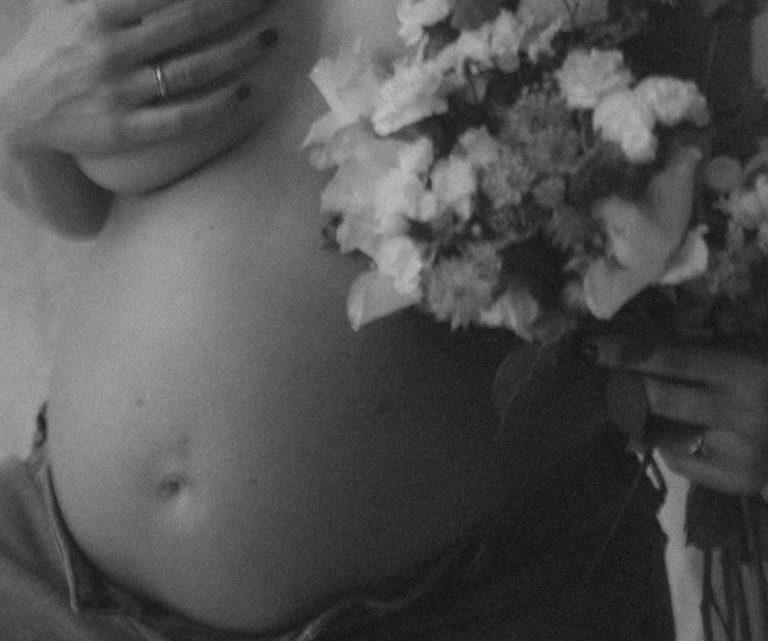 Schwangere Person mit nacktem Oberkörper, im Fokus der Bauch im 10. Schwangerschaftsmonat, der Busen durch einen Blumenstrauss verdeckt.
